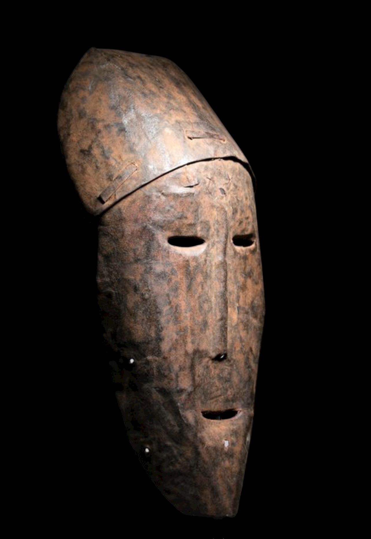 Een hoog, verweerd, donkerbruin houten masker met een langwerpig gezicht en een opvallend uitstekend voorhoofdsstuk.