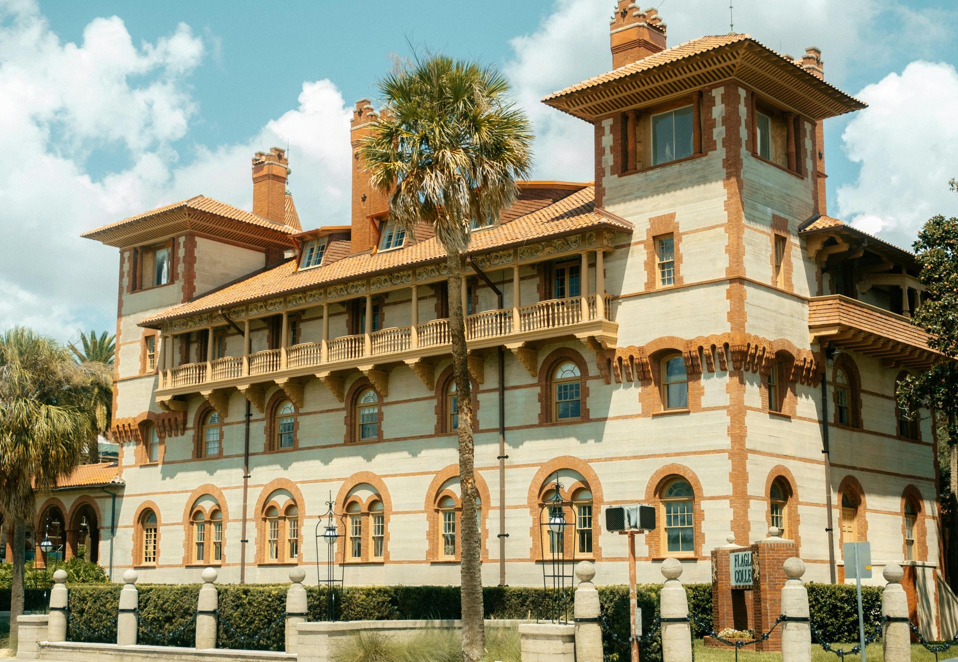 Historic beige mansion with balconies and red-tiled roofs, surrounded by palm trees under a blue sky
