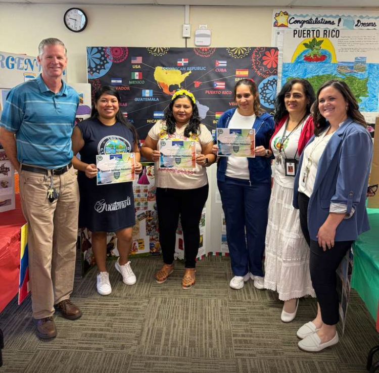 Group of people holding certificates in a classroom with a world map and flags.