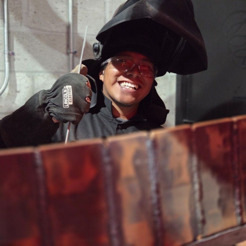 Person welding, smiling, wearing protective gear, indoors.