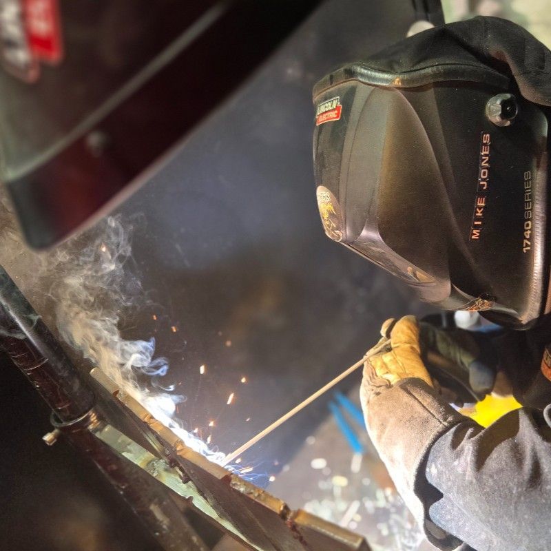 Person welding, wearing a protective helmet, gloves, and jacket, with sparks and smoke in a workshop.