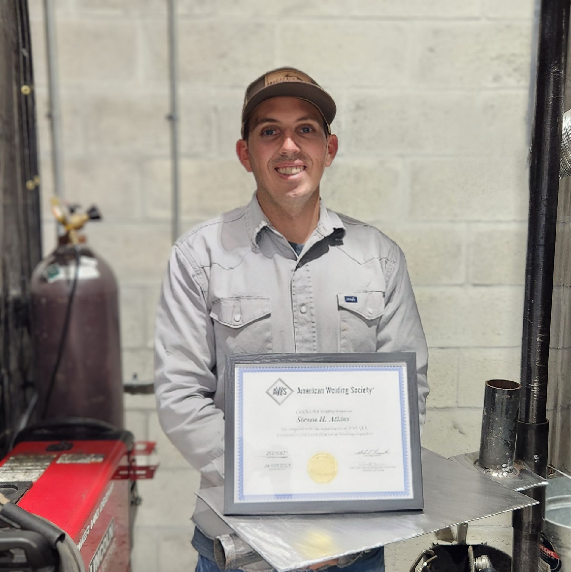 Man in work clothes holding a framed American Welding Society certificate in a workshop.