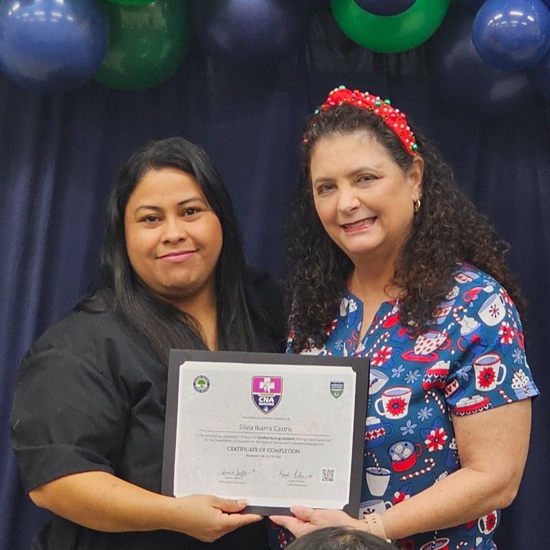 Two women holding a framed certificate; one wears a festive headband and scrub top.