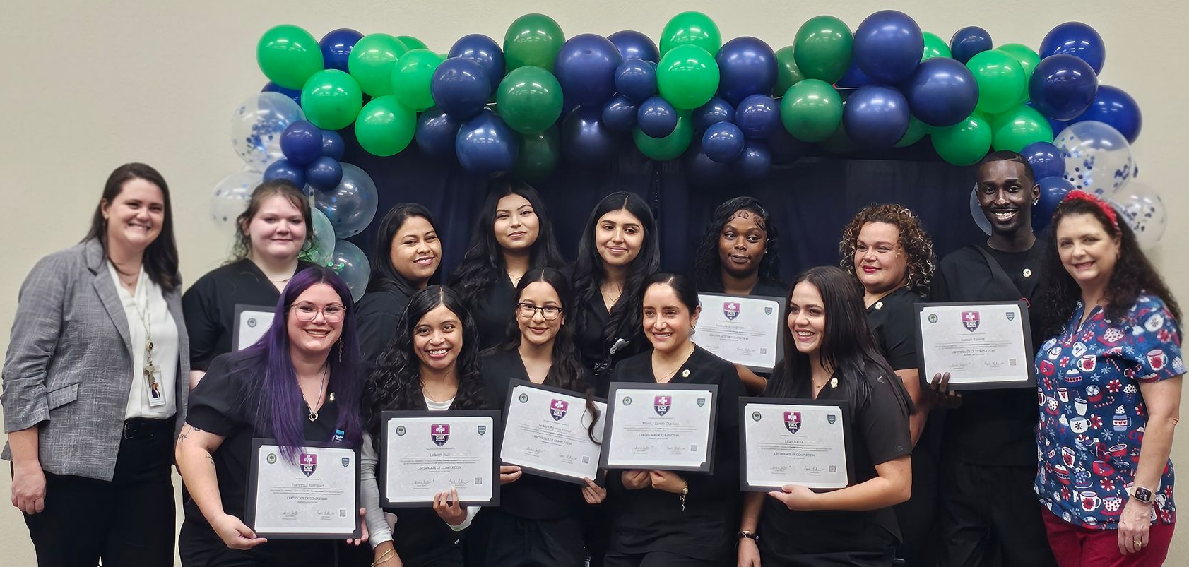 Group of people in scrubs holding certificates in front of balloons.