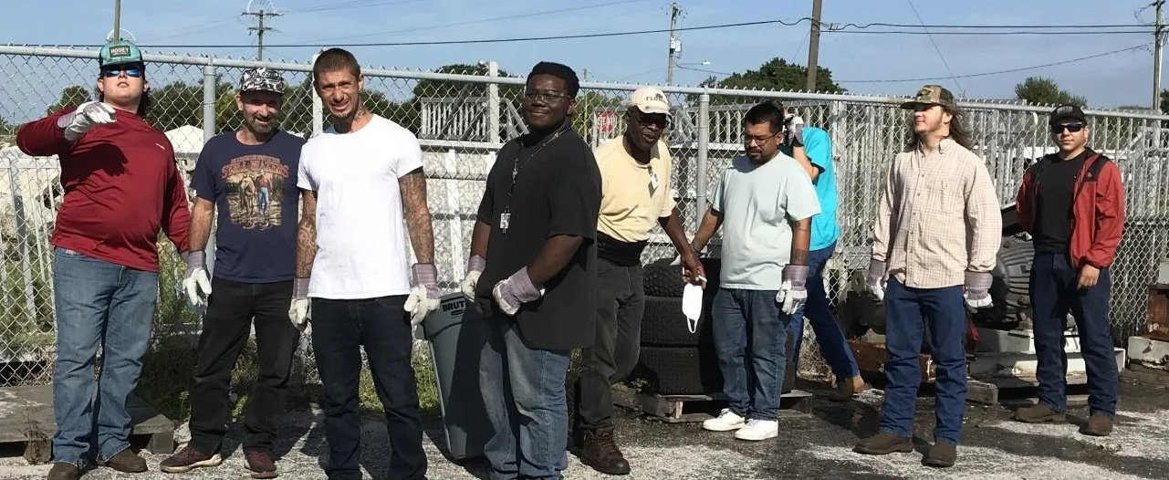 Group of people standing outside, possibly working. Some wear gloves, and a chain link fence is behind them.