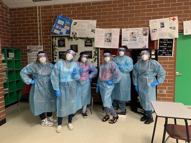 Six people in protective gear pose in a classroom, wearing face shields, gowns, and gloves.