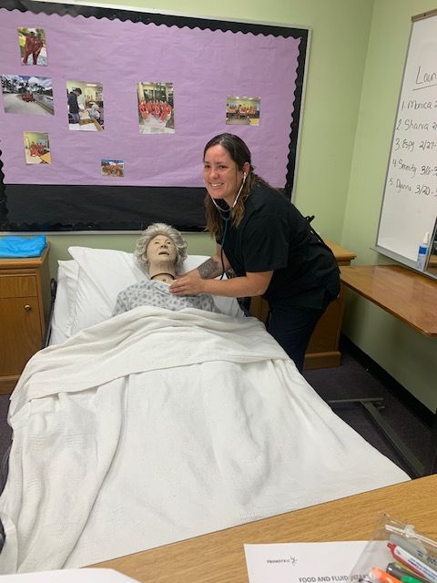 Woman using stethoscope on a mannequin in a classroom. The woman is smiling.