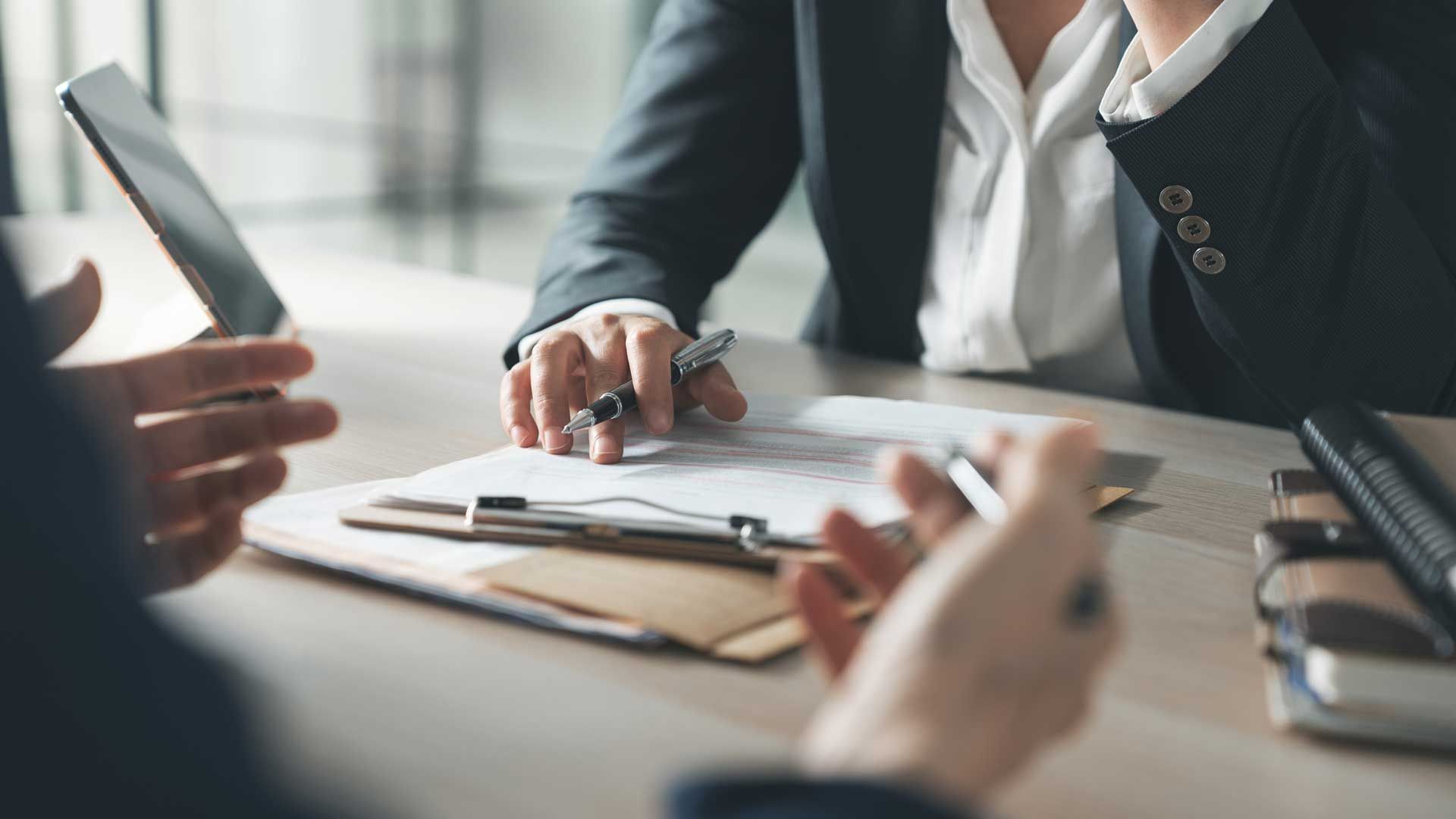 Business meeting with hands over a desk, papers, tablet, and pen in a bright office.