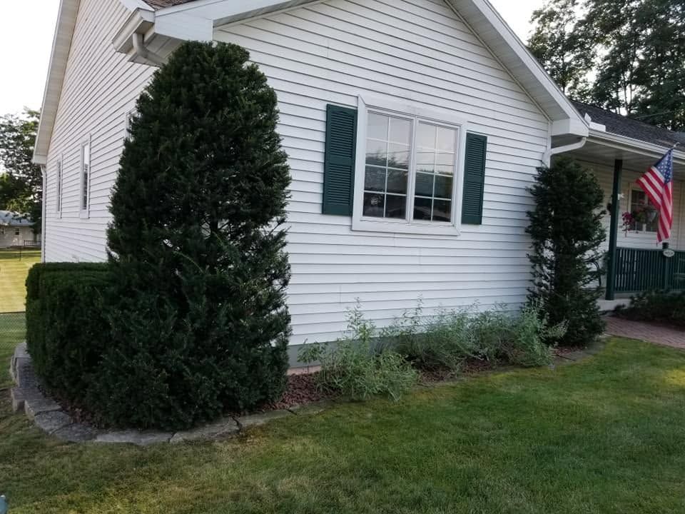 White house exterior with green shutters, trimmed shrub, flower bed, and an American flag by the porch