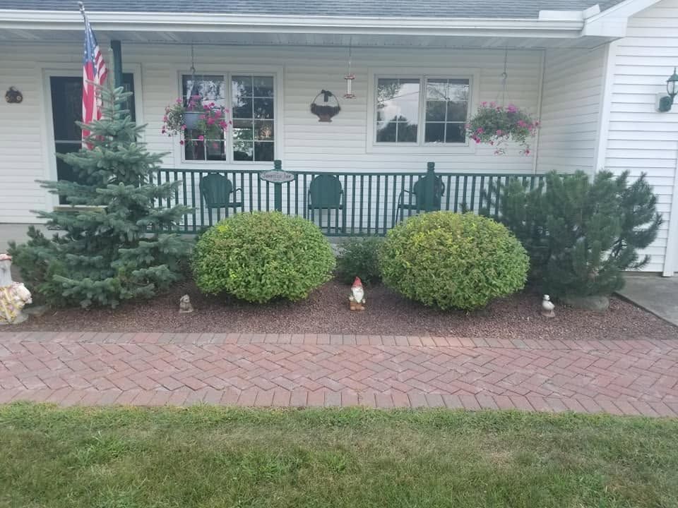 Front porch with white siding, black railing, potted plants, shrubs, brick walkway, and an American flag