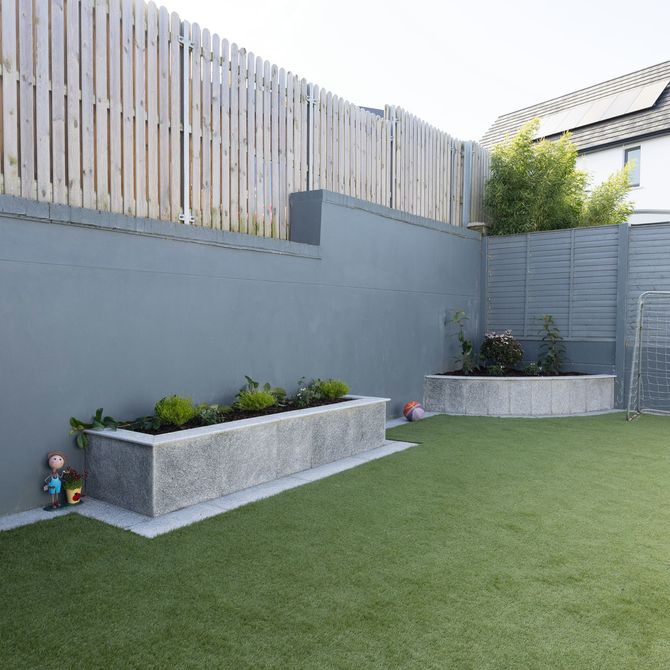 A backyard with green artificial turf, a smooth grey wall, a wooden fence, and two stone planters filled with shrubs.