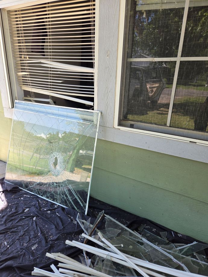 Broken window with a cracked pane leaning against a house exterior below two windows and scattered glass shards