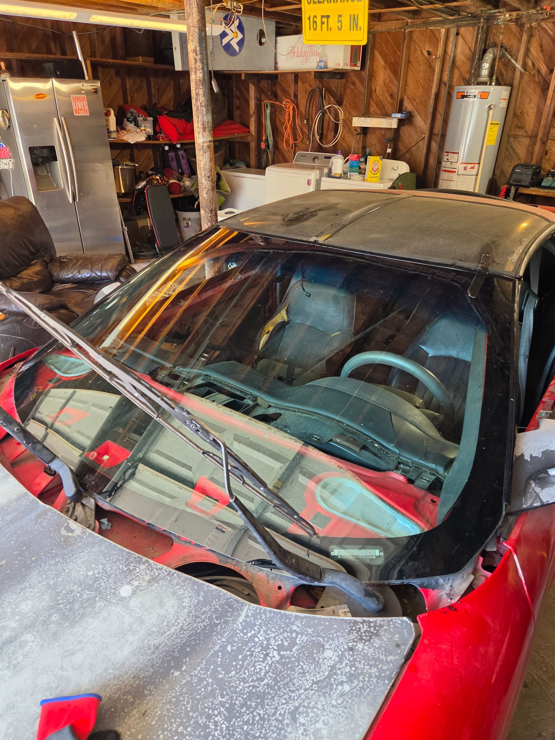 Red sports car parked in a cluttered garage, with tools and storage shelves around it.