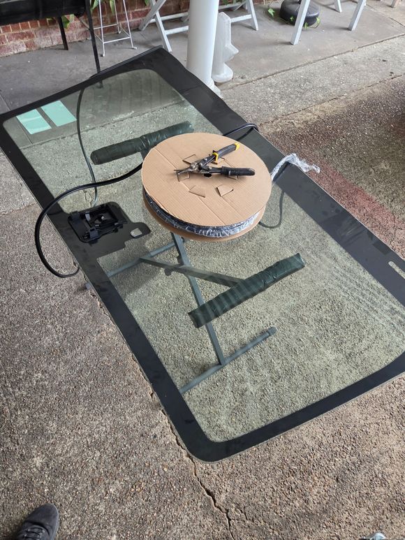 Outdoor glass patio table with a round wooden board, small tools, and a black smartphone on a speckled patio floor
