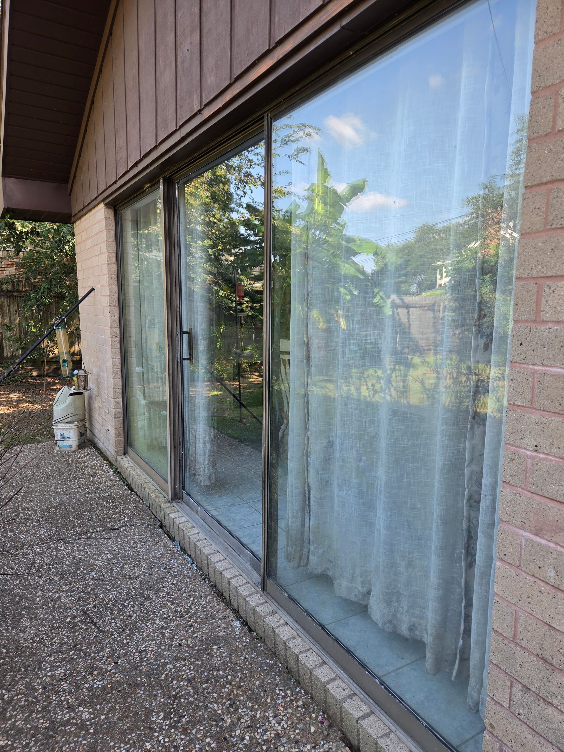 Exterior view of large sliding glass doors with curtains, reflecting trees and blue sky beside a gravel patio