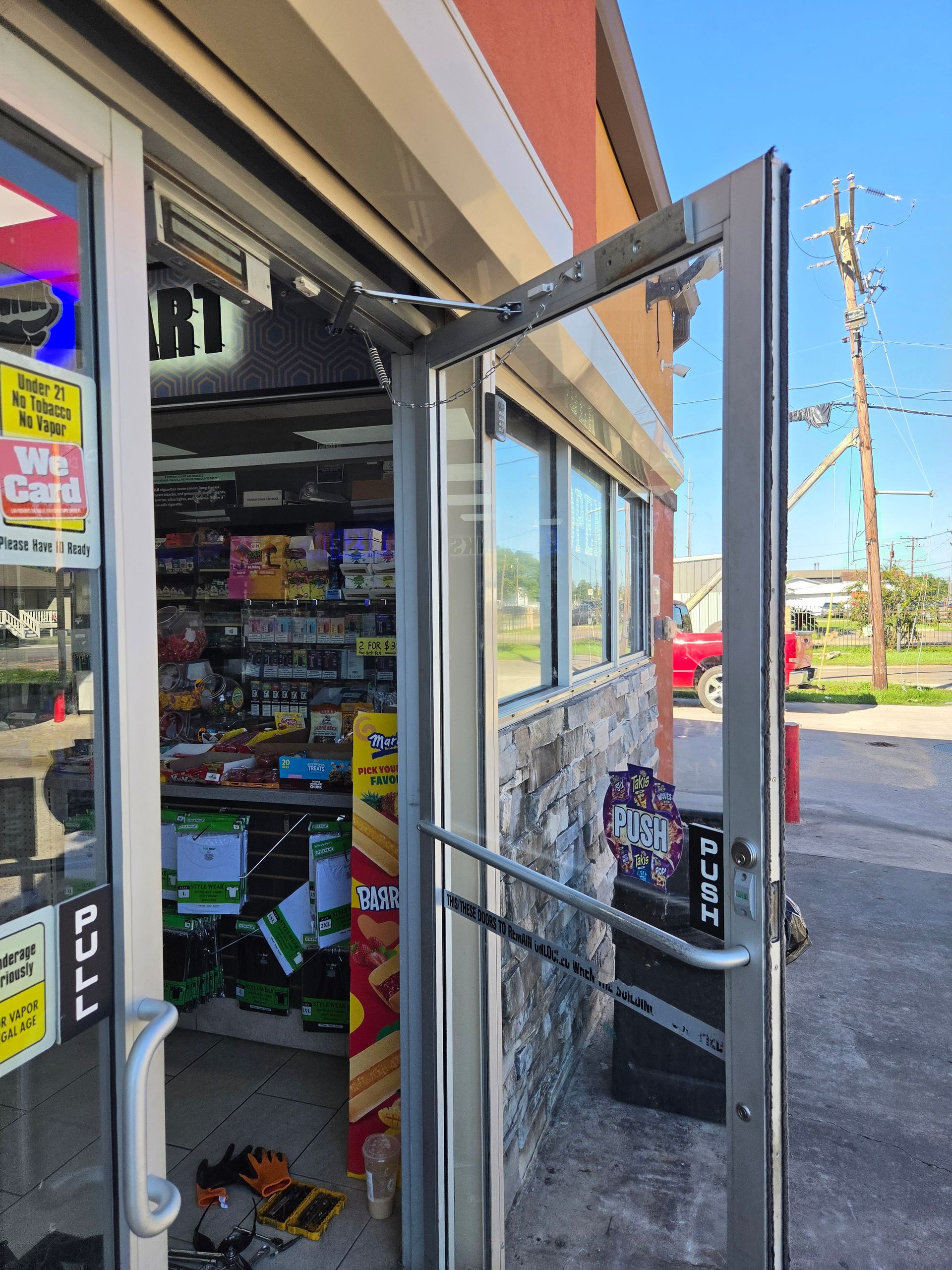 Open storefront door with shelves of snacks and drinks visible inside, sunny sidewalk outside