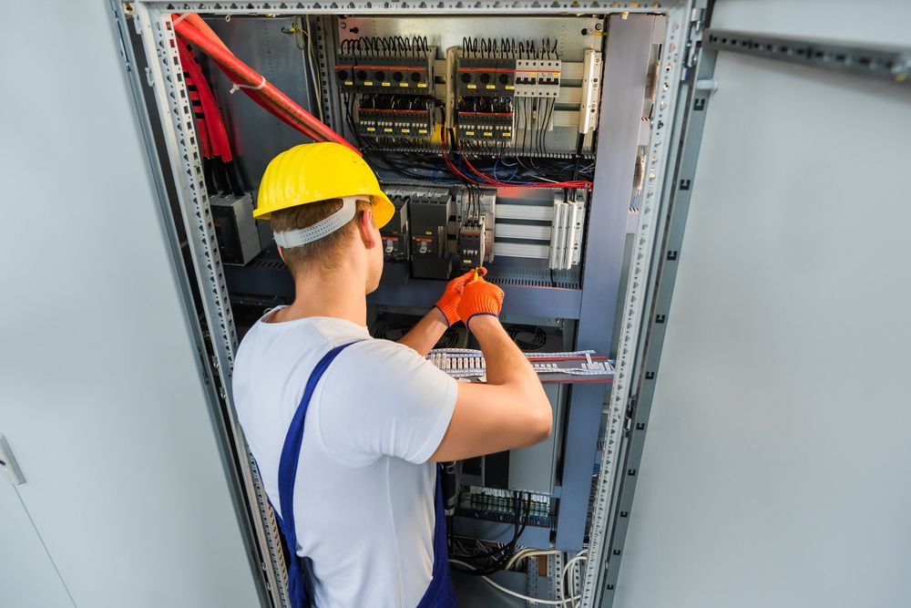 Electrician in yellow hard hat and orange gloves works on electrical panel.