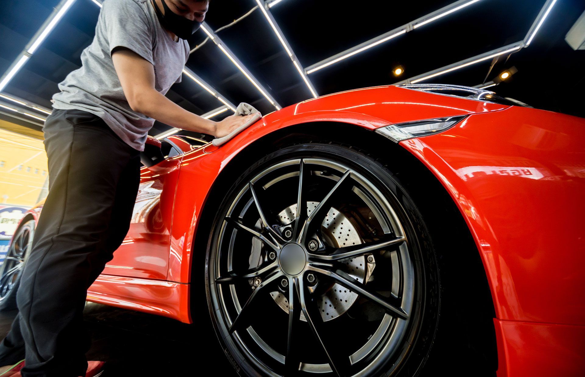 Person in mask polishing a bright red sports car wheel in a brightly lit garage.