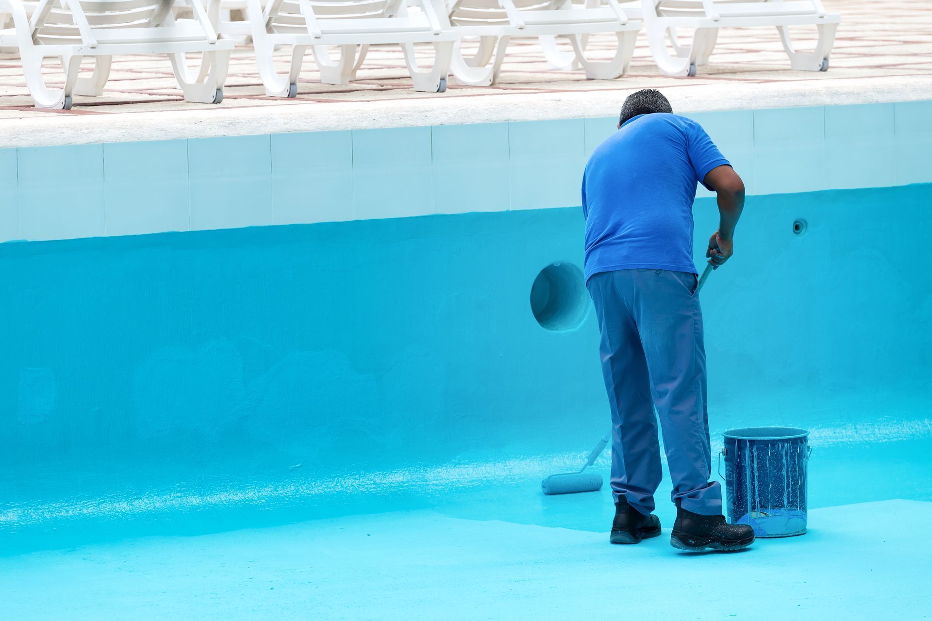 Worker cleaning an empty turquoise pool with a hose and bucket.