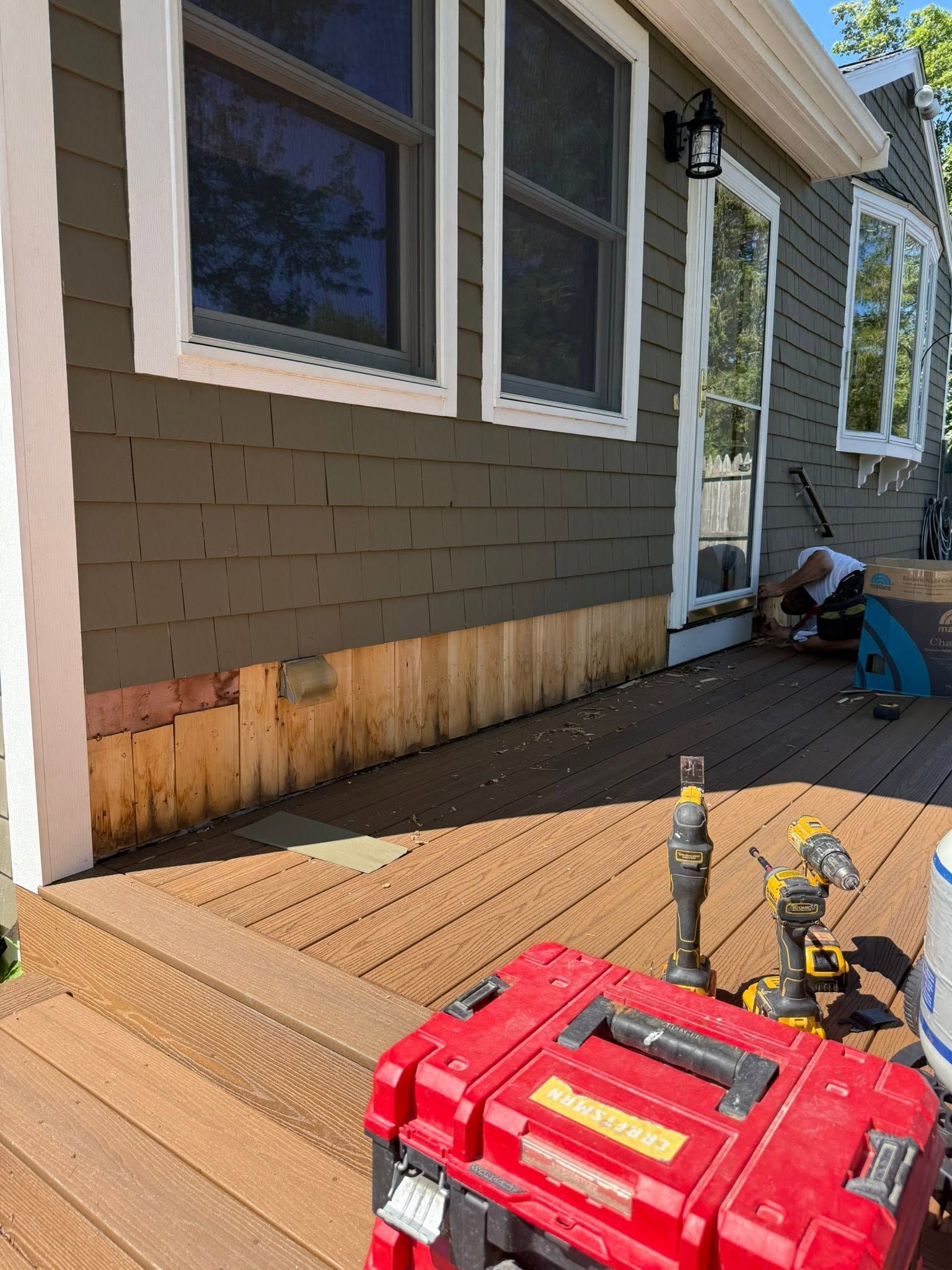 Deck repair work beside a house, with tools and exposed damaged siding near the wall.