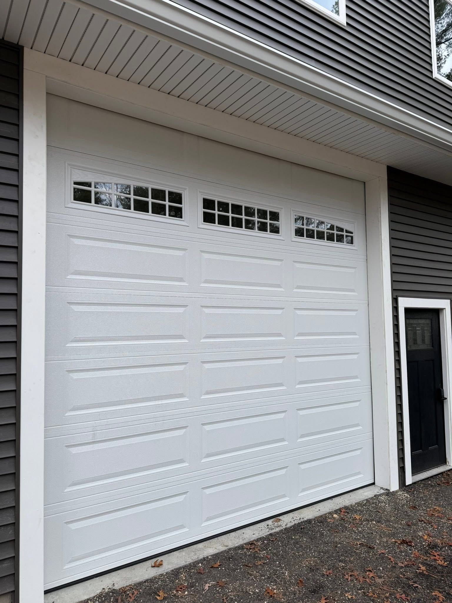 White garage door with small windows beneath a house overhang, beside a black side door.