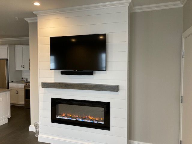 A wall-mounted TV and linear electric fireplace installed on a white shiplap feature wall with a dark wood mantel.