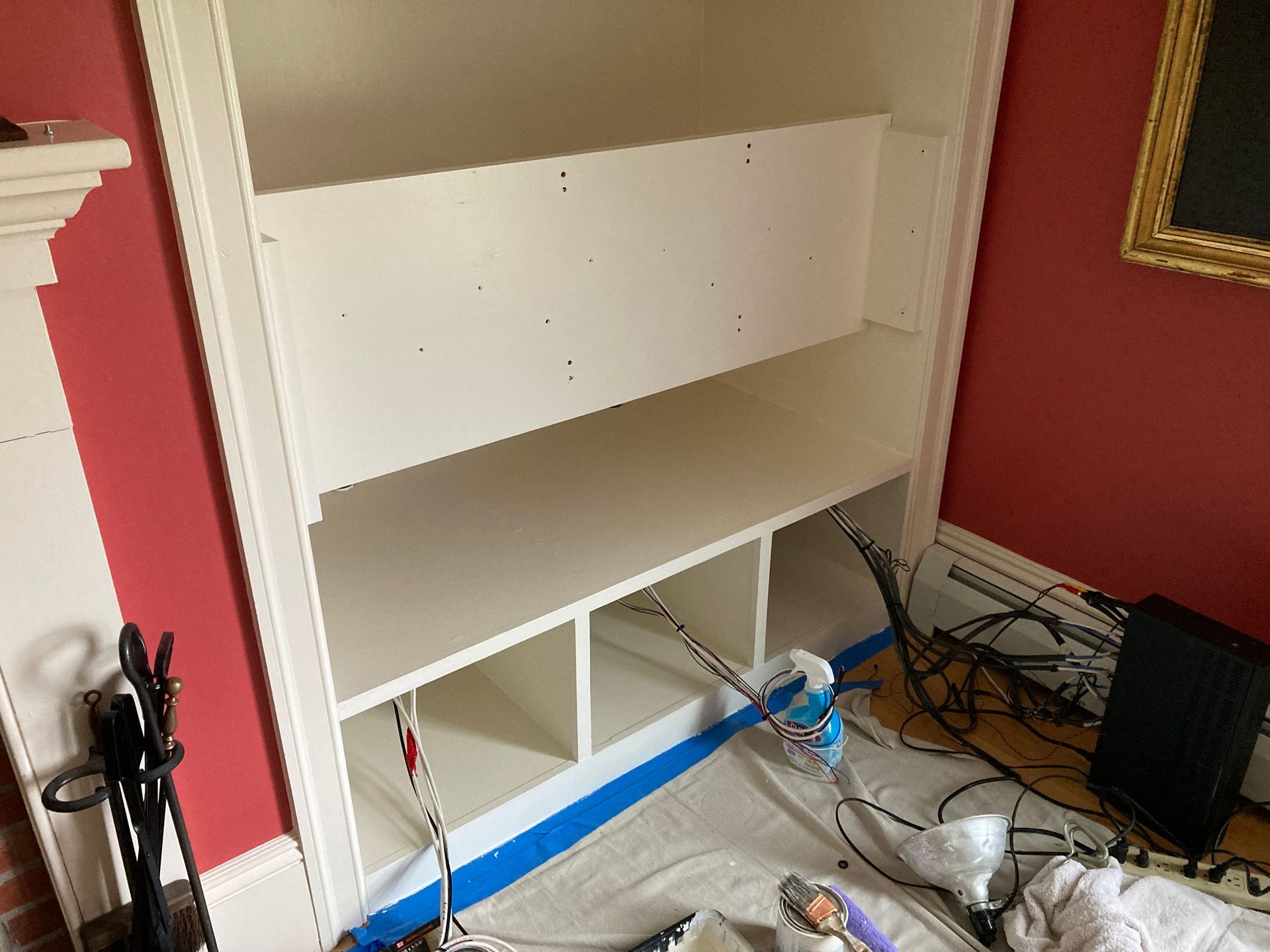 A white, partially constructed built-in bookshelf set against a red wall, with visible wires and equipment on the floor.