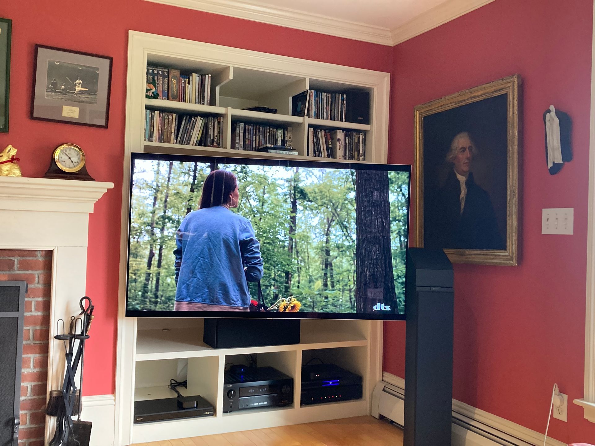 A television showing an outdoor forest scene sits in a built-in white shelf against a red wall with framed portraits.