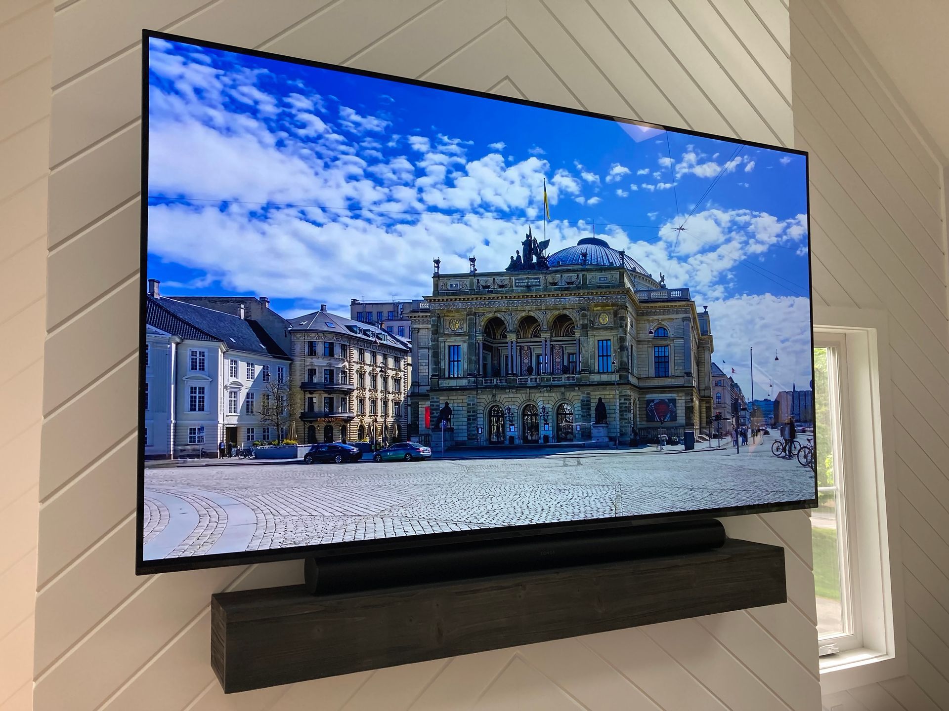A flat-screen TV mounted on a white, chevron-patterned wall, displaying an image of a classical building in a plaza.