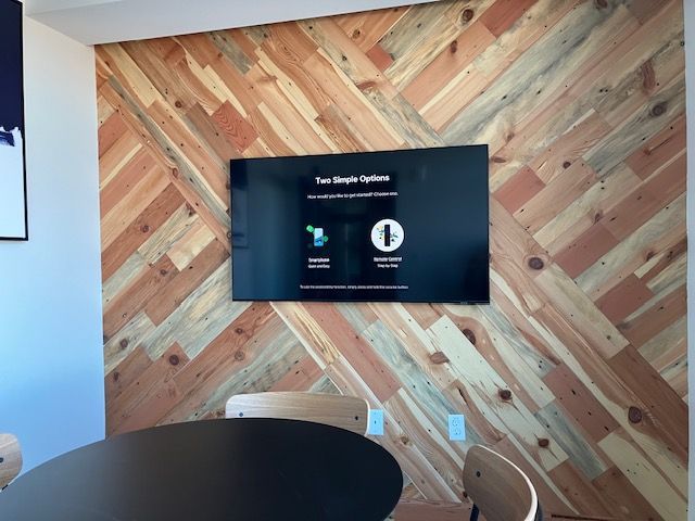A flat-screen TV mounted on a wall with diagonal wood paneling in a room with a round table and chairs.