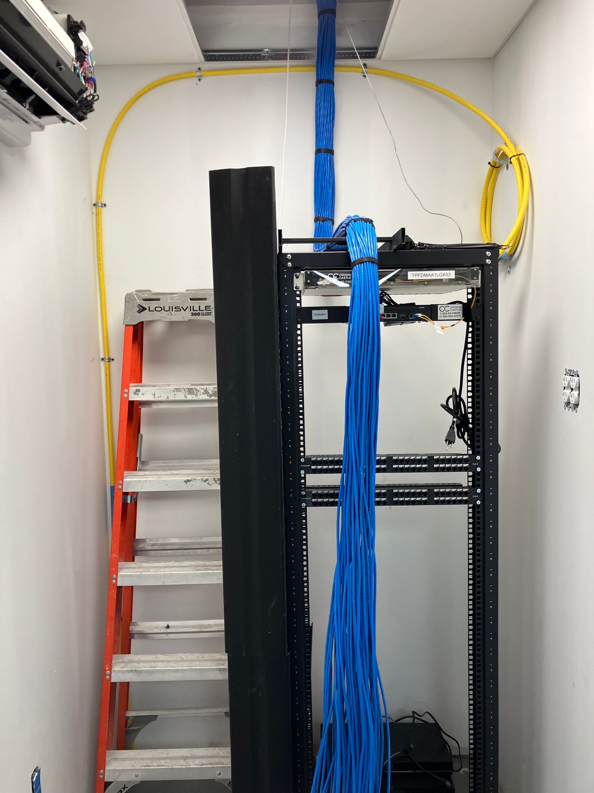 A network server rack with hanging blue cables, positioned next to an orange ladder in a small white server room.