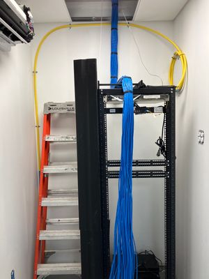 A network server rack with hanging blue cables, positioned next to an orange ladder in a small white server room.