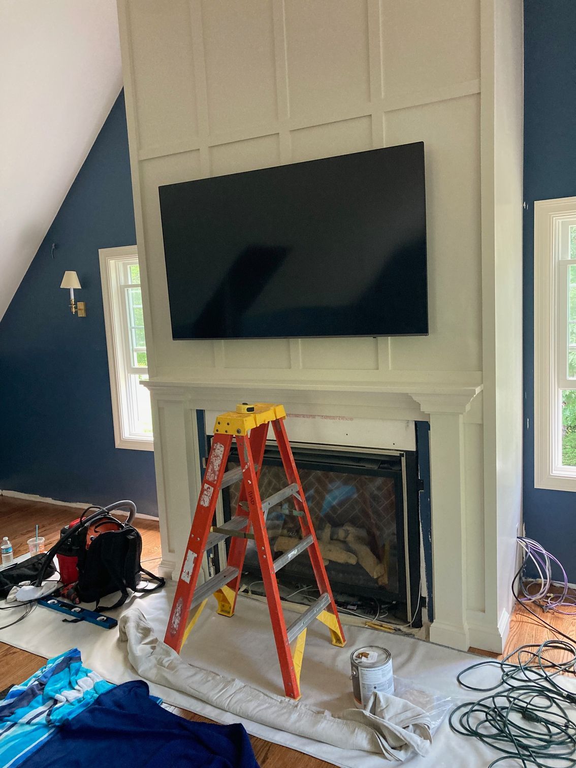 A TV mounted above a fireplace, with a red ladder and construction supplies on a drop cloth in a blue-walled room.