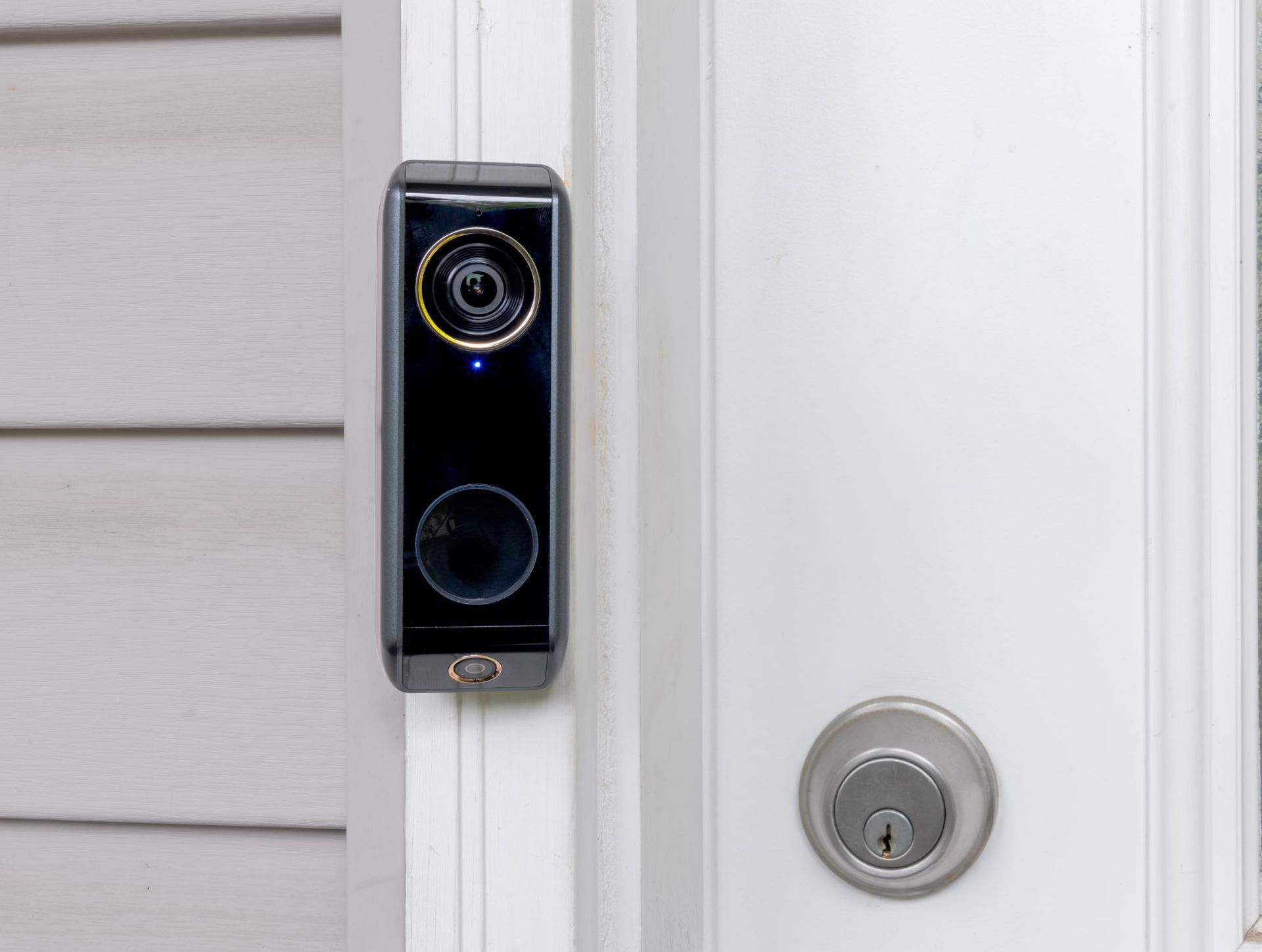 A black video doorbell mounted on a white door frame next to a white door with a silver deadbolt.