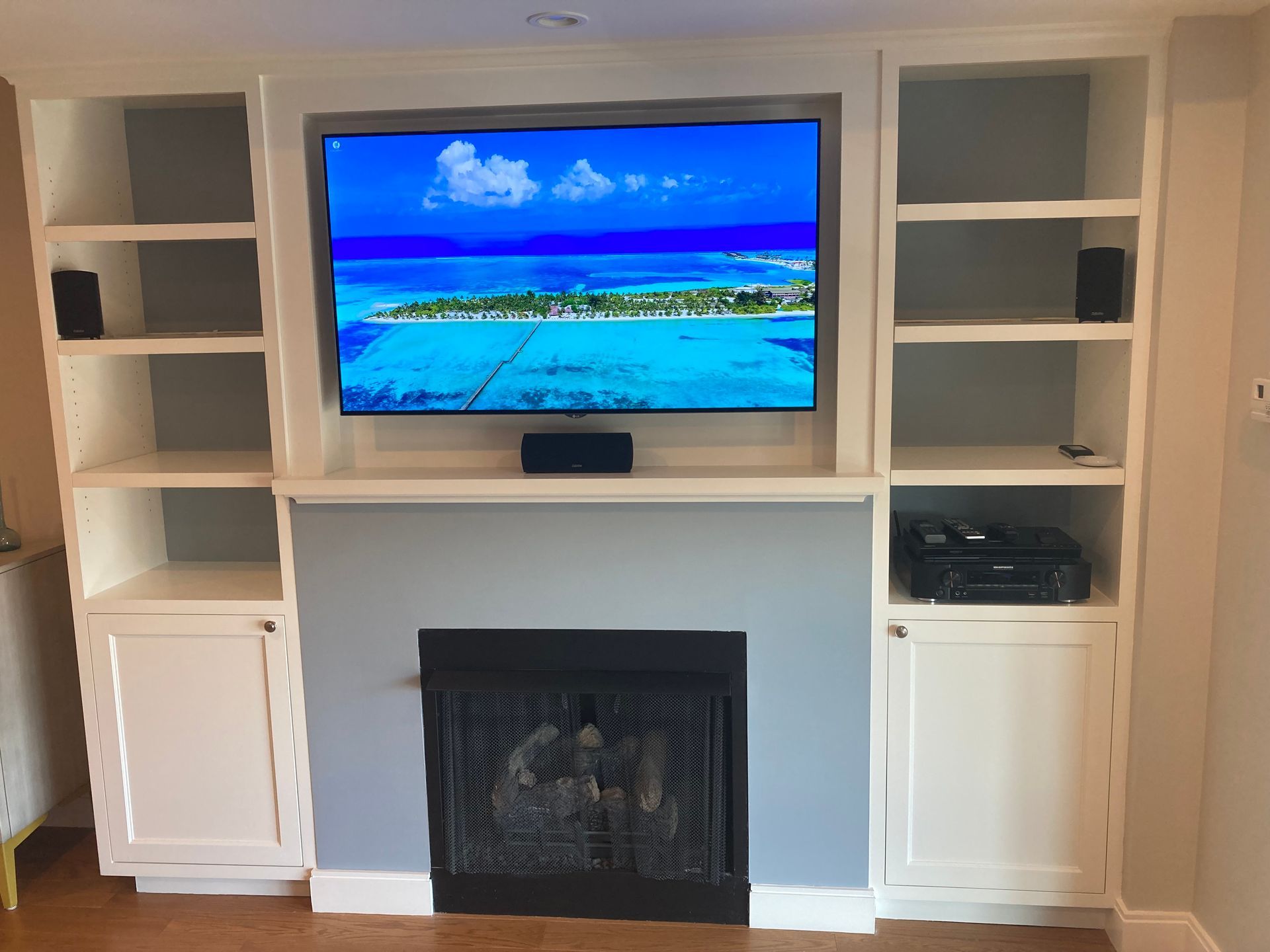 A wall-mounted TV above a fireplace, flanked by white built-in shelving units against a light gray wall.