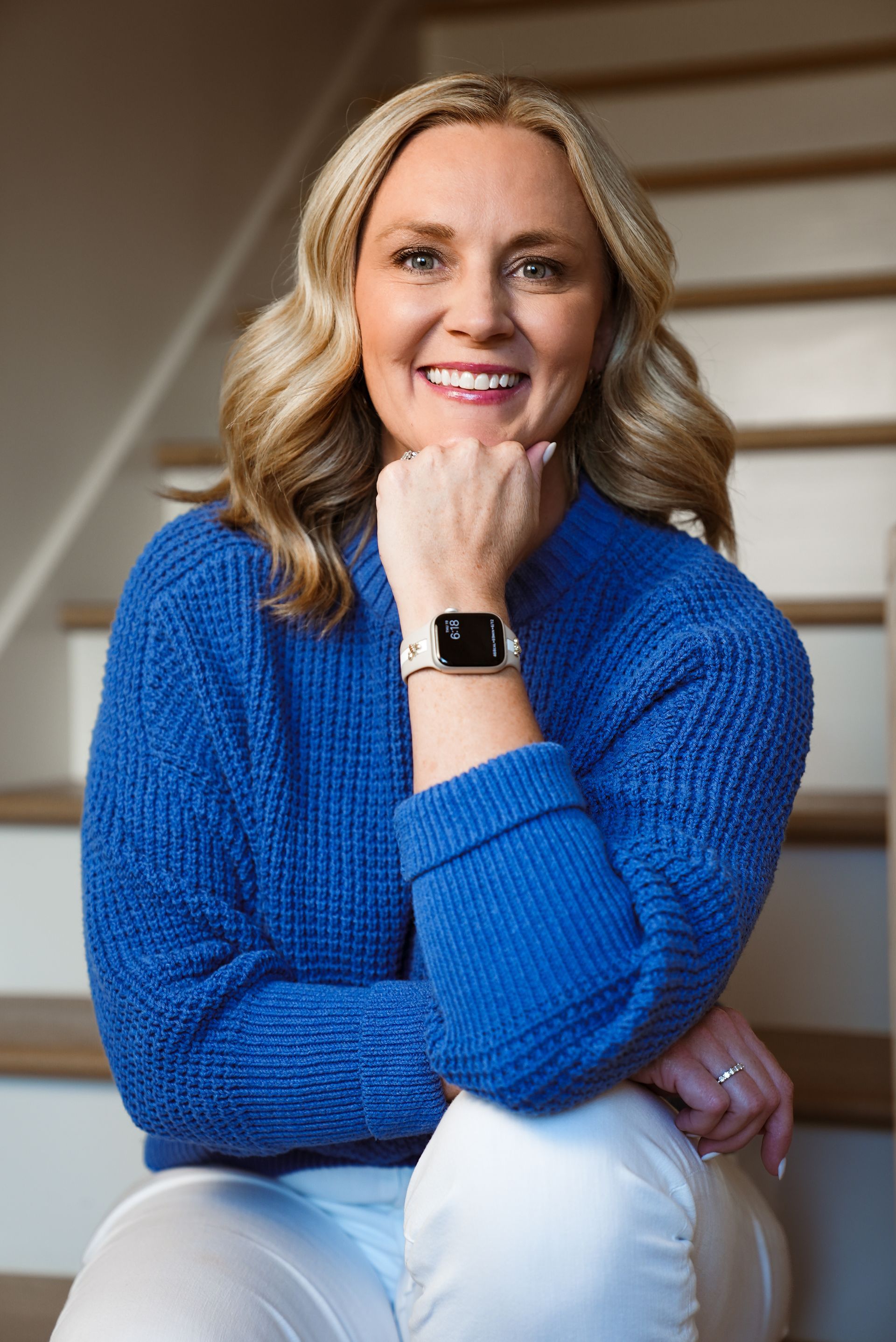 A woman in a blue sweater rests her chin on her fist, smiling into the camera for a professional headshot.