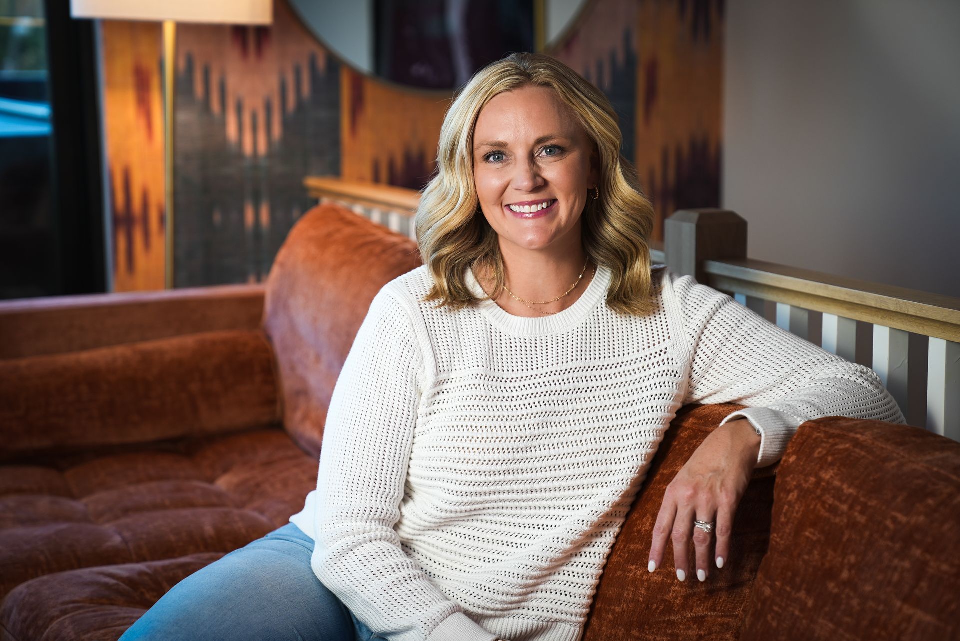 A smiling woman in a white sweater leans back against a brown couch for a professional headshot.