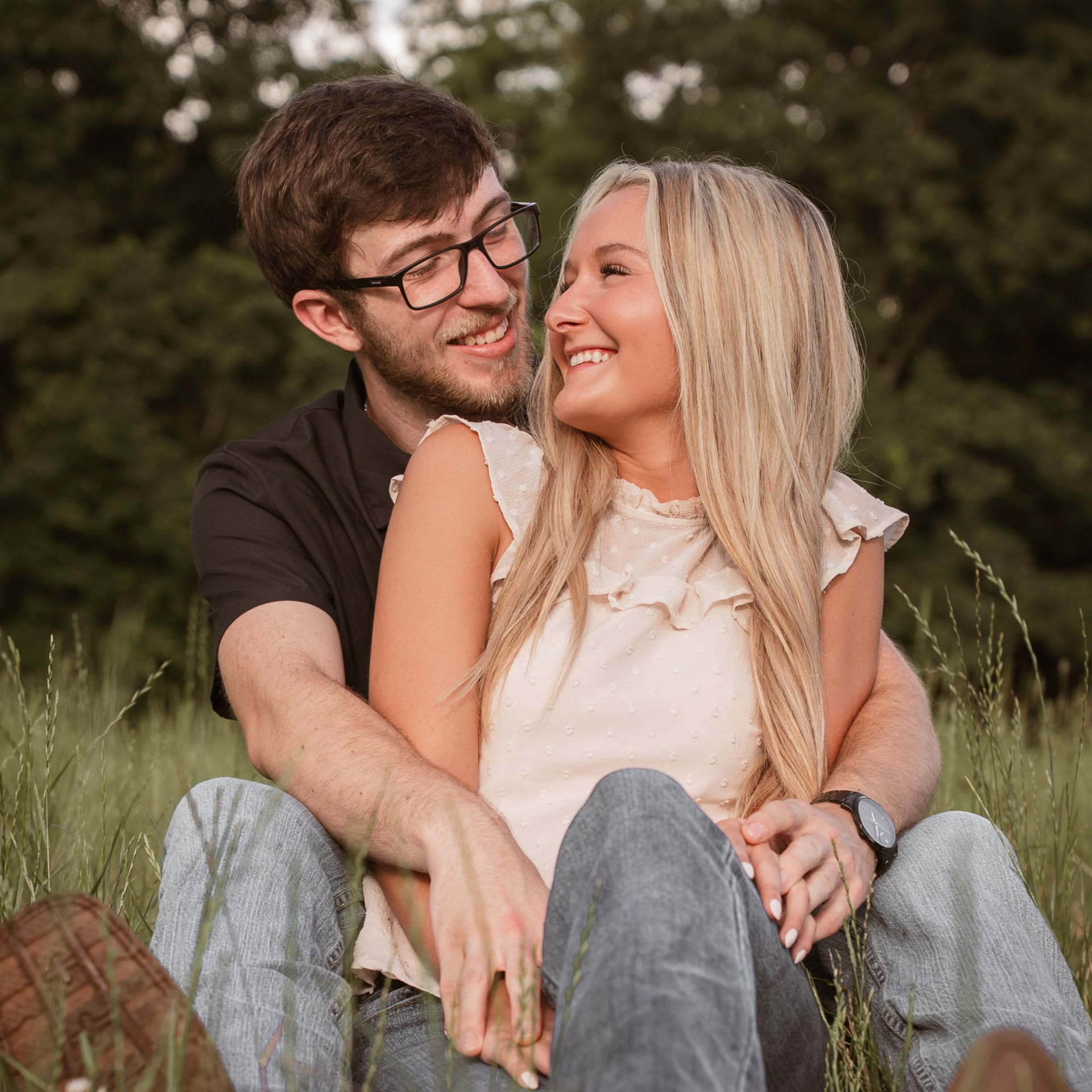A couple sit in a field, holding hands and smiling at each other.