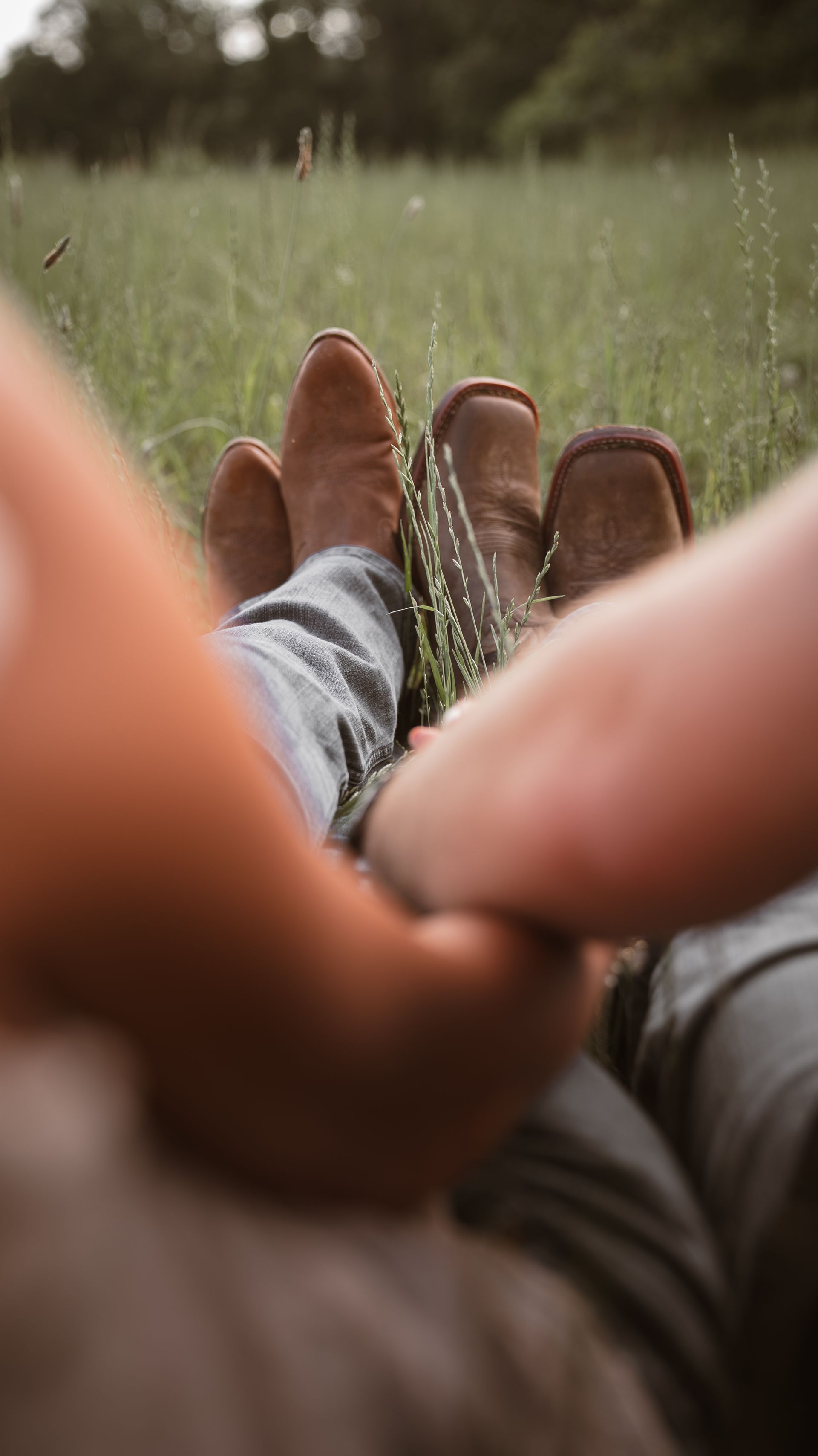 Close up of a couple sitting in a field wearing cowboy boots and holding hands.