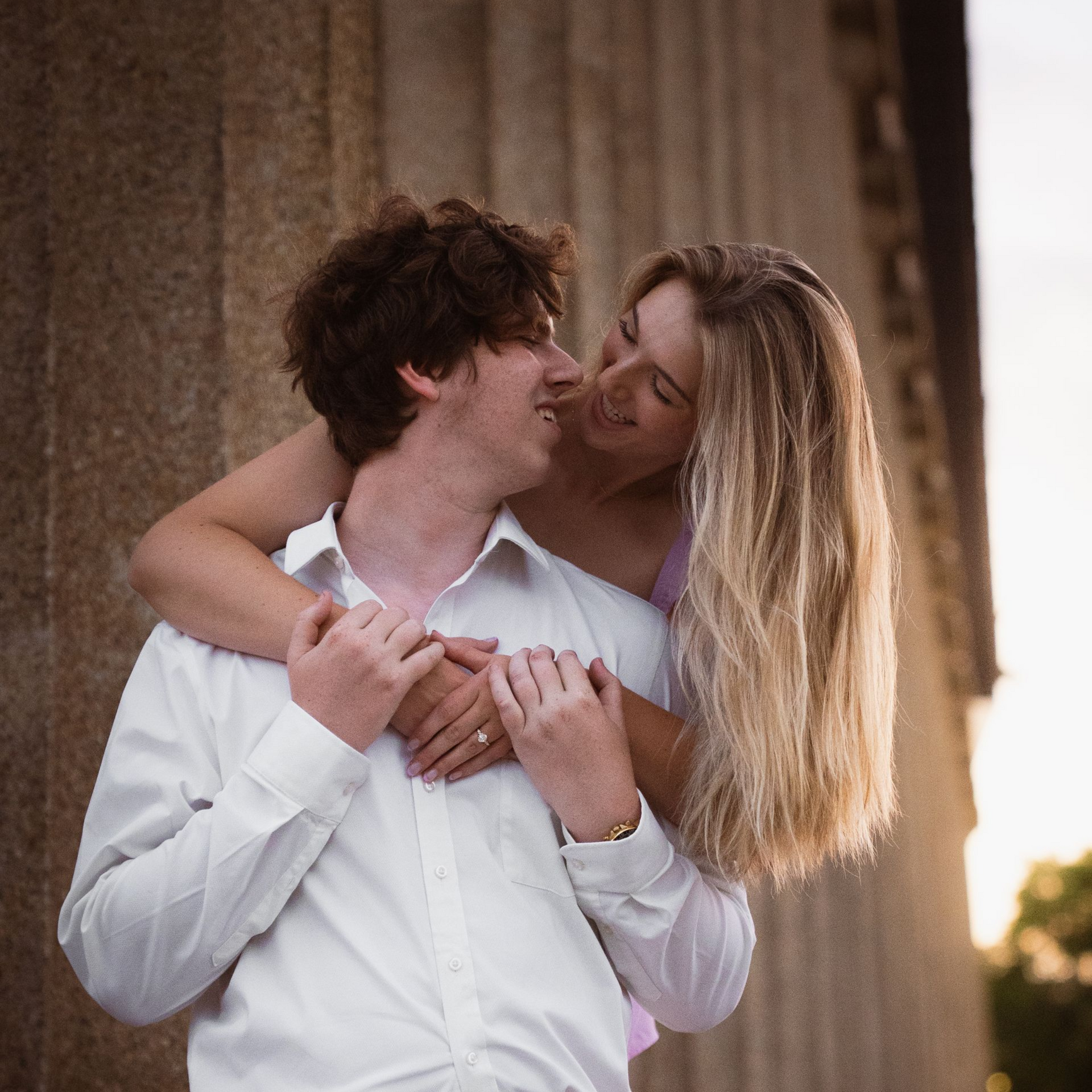 A woman leans over her fiancee's shoulder at sunset, smiling at him.
