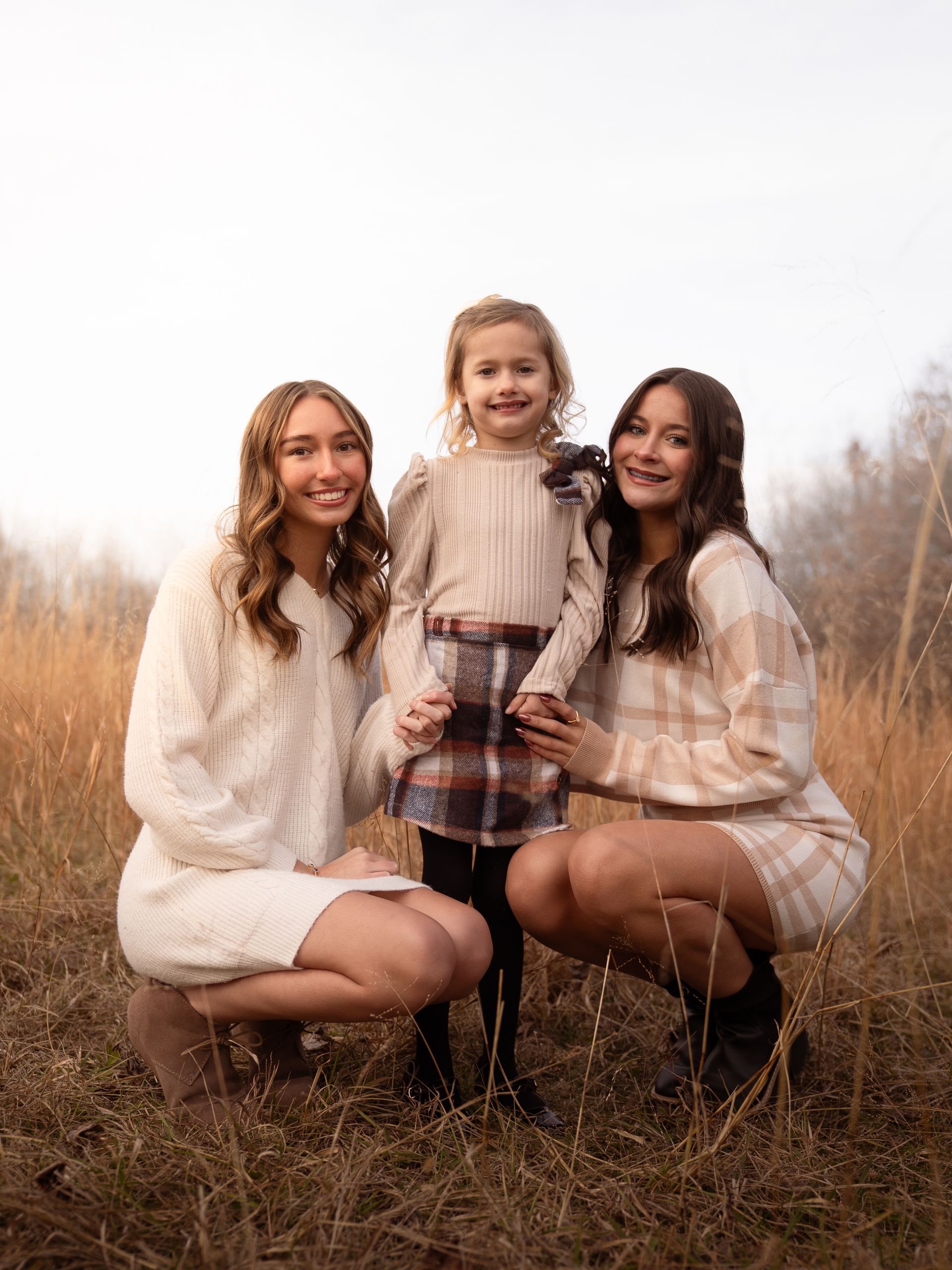Two girls hold their younger cousin between them in a field at sunset, posing for family photos.