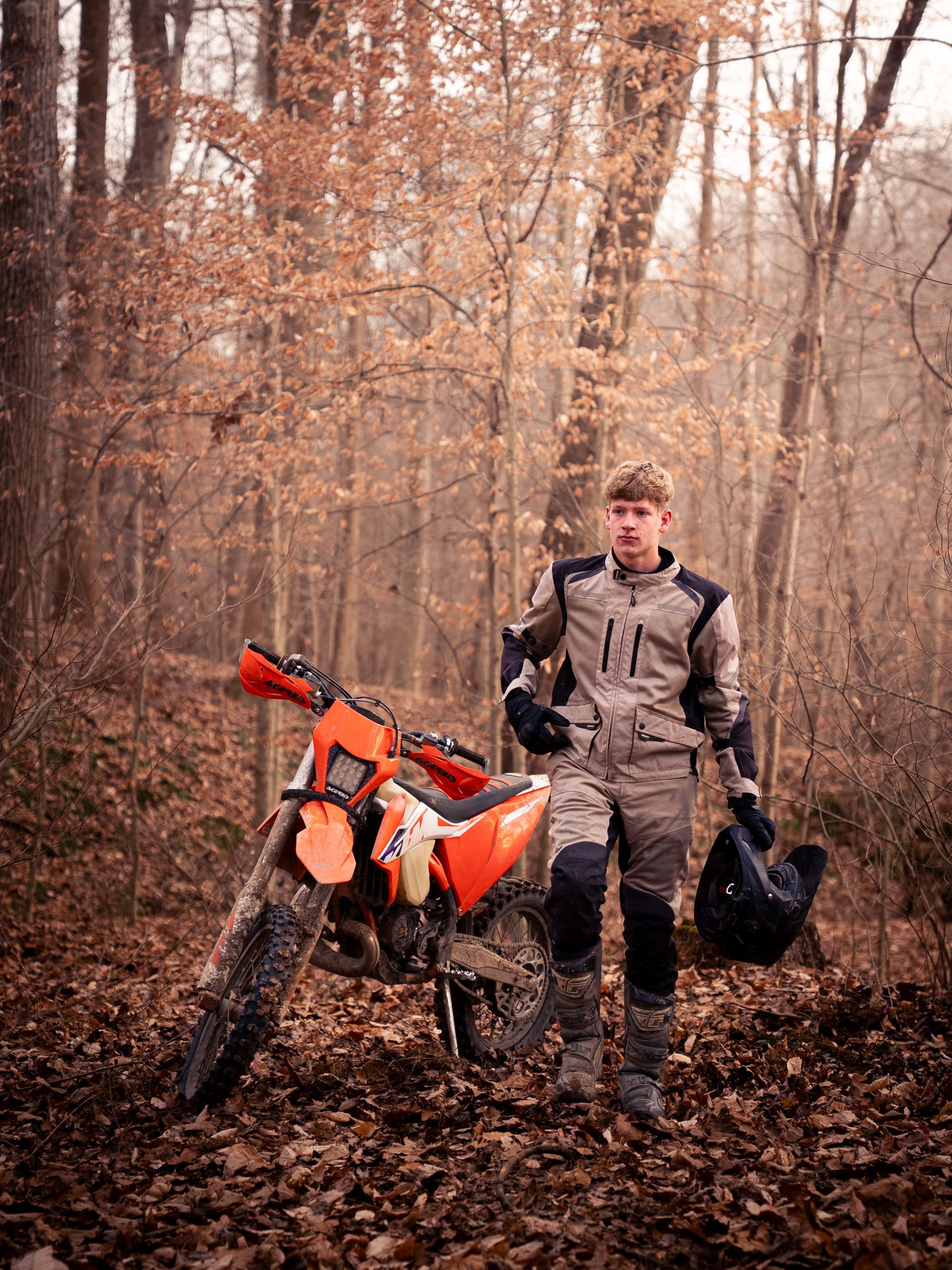 Boy walking through the woods next to his dirt bike holding his helmet for senior photos.
