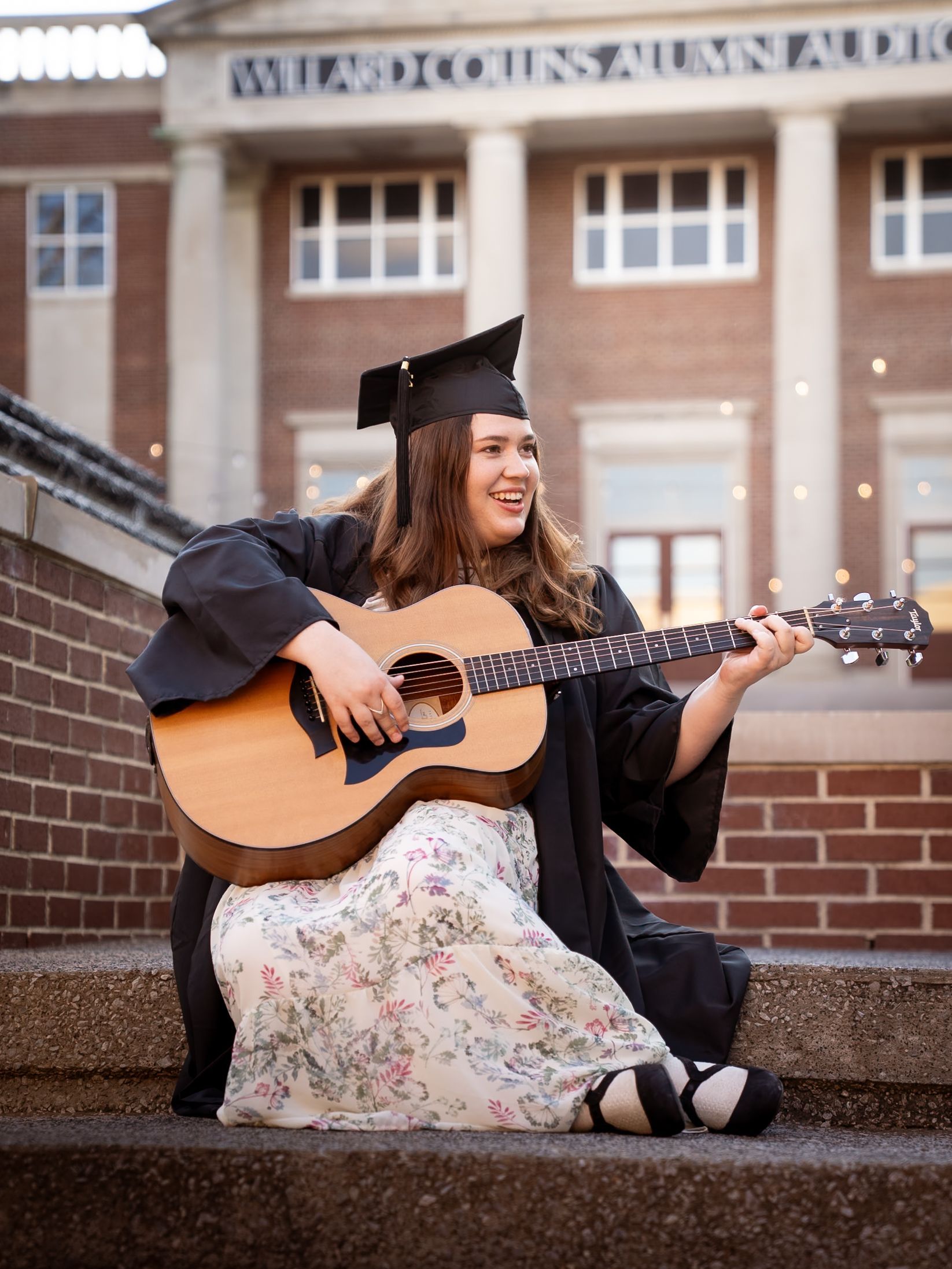 Woman wearing a graduation cap and gown sitting next a fountain playing the guitar.
