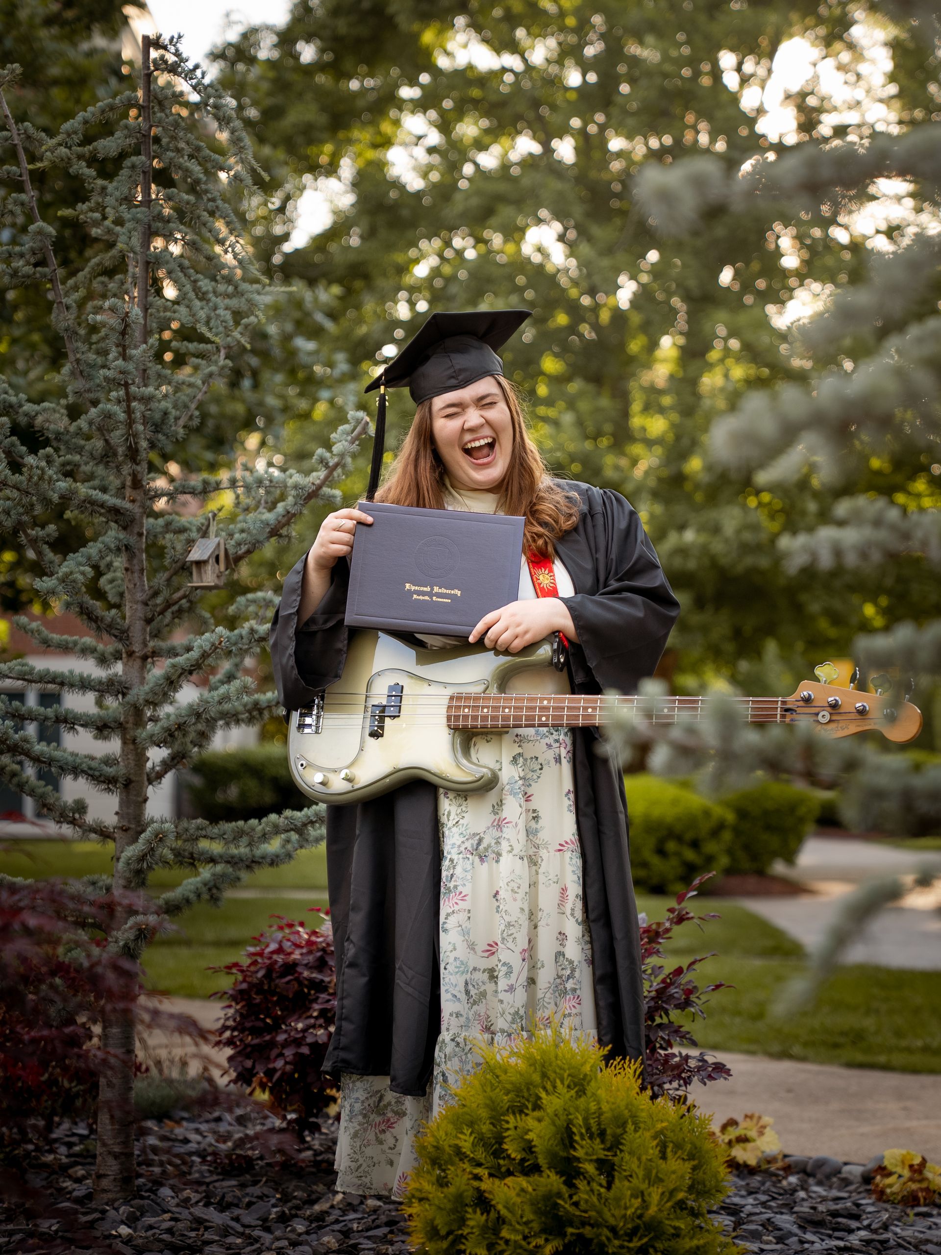 Woman standing in greenery wearing a graduation cap and gown and holding her diploma.