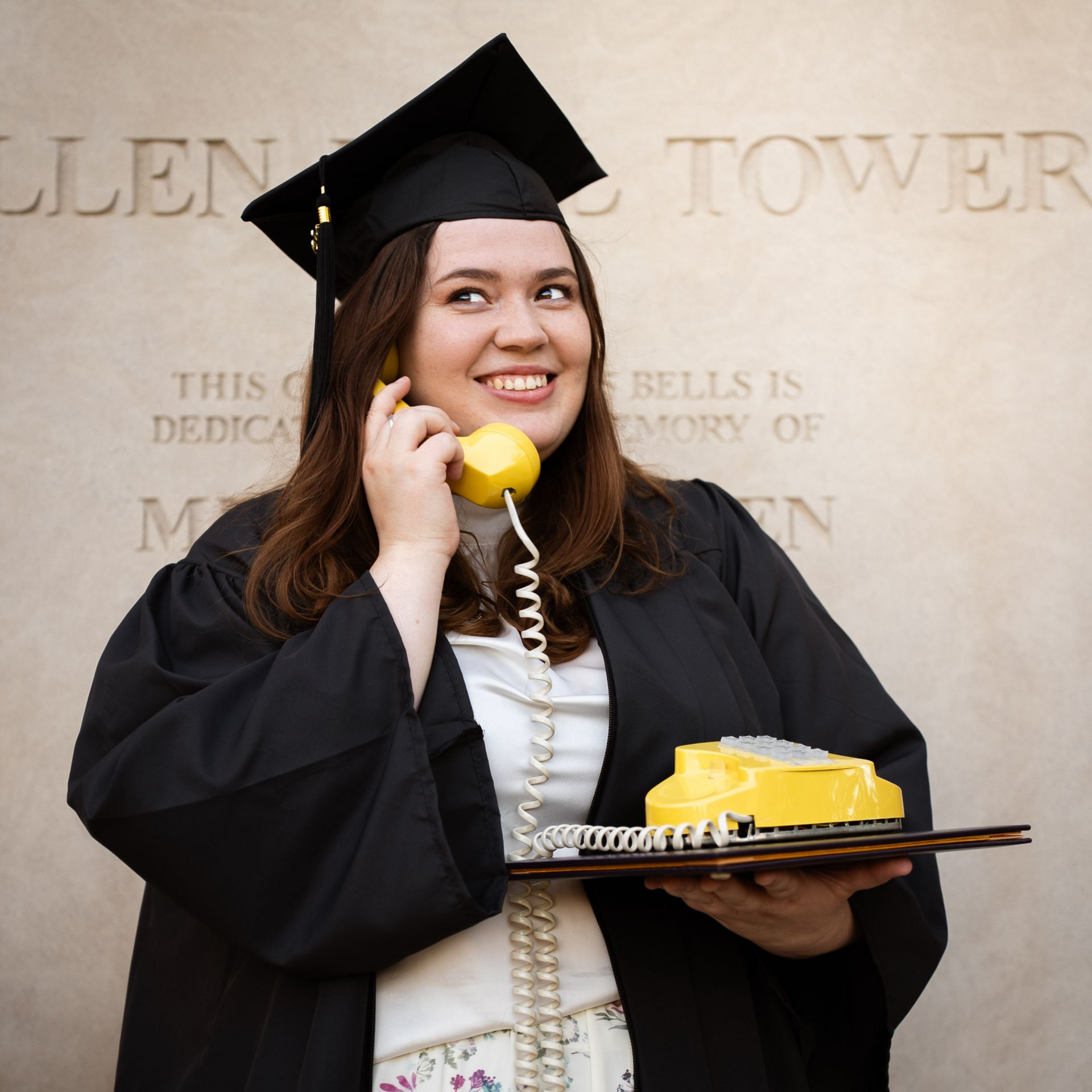 Woman holding an old yellow phone with a graduation cap and gown on for graduation photos.