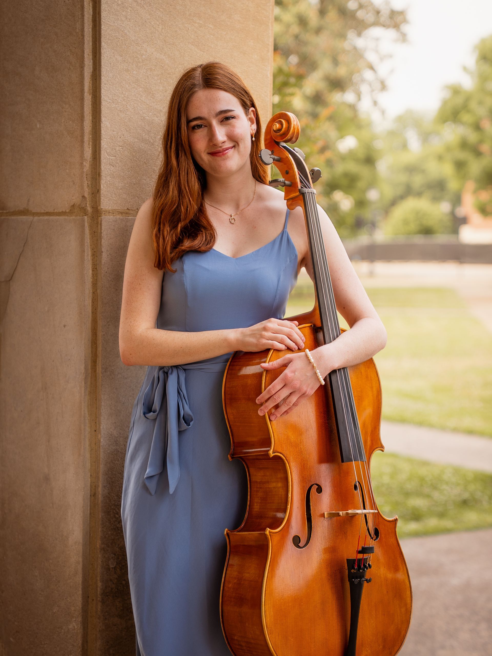 Woman standing against a column holding a cello at sunset for senior photos.