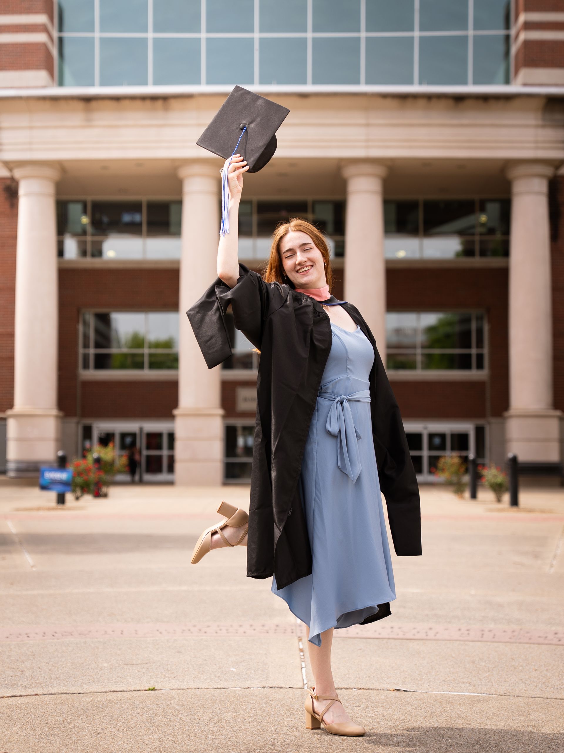 Woman wearing a graduation gown standing in front of a library holding her cap in the air.