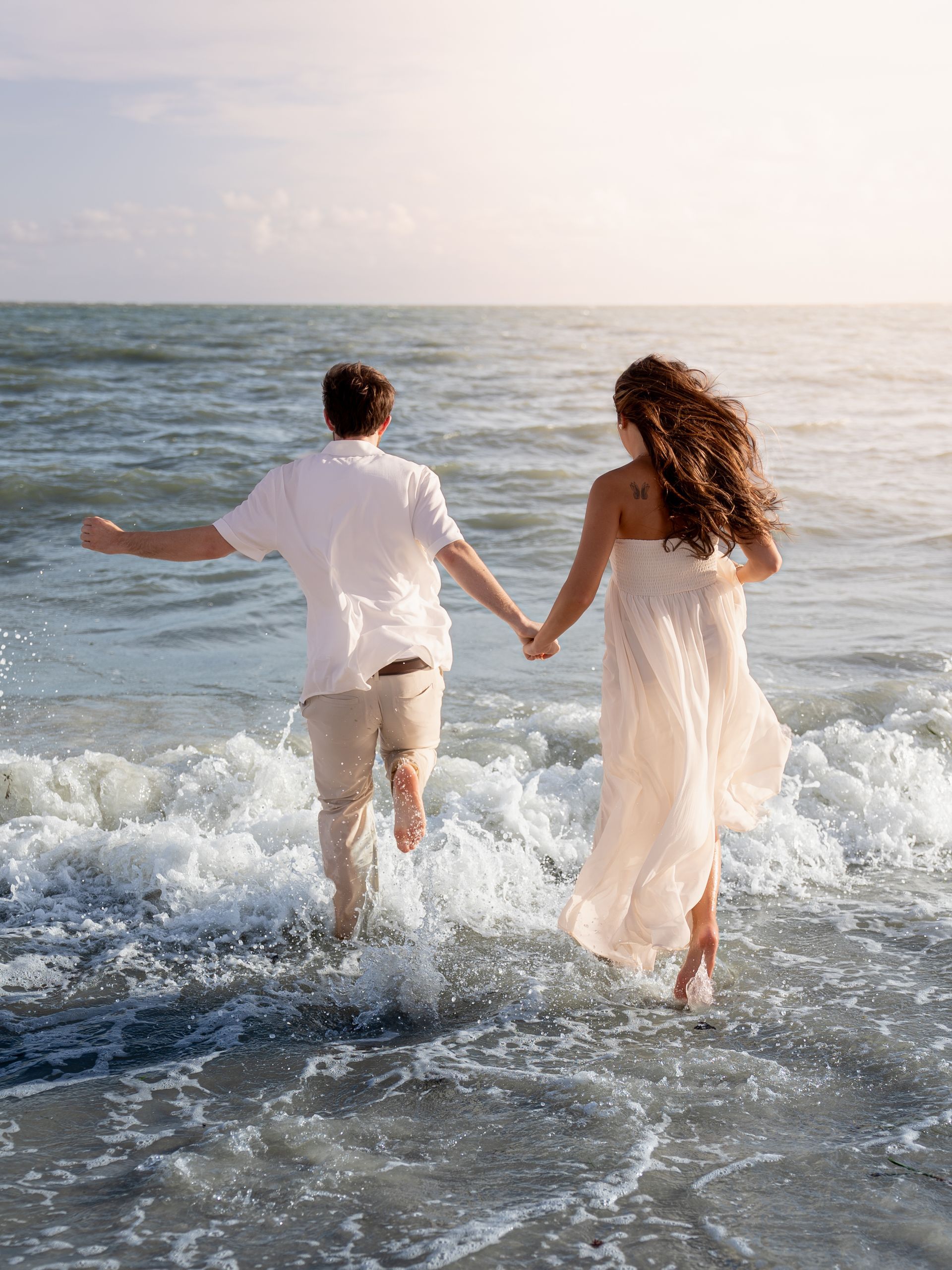 A couple runs into the ocean holding hands.
