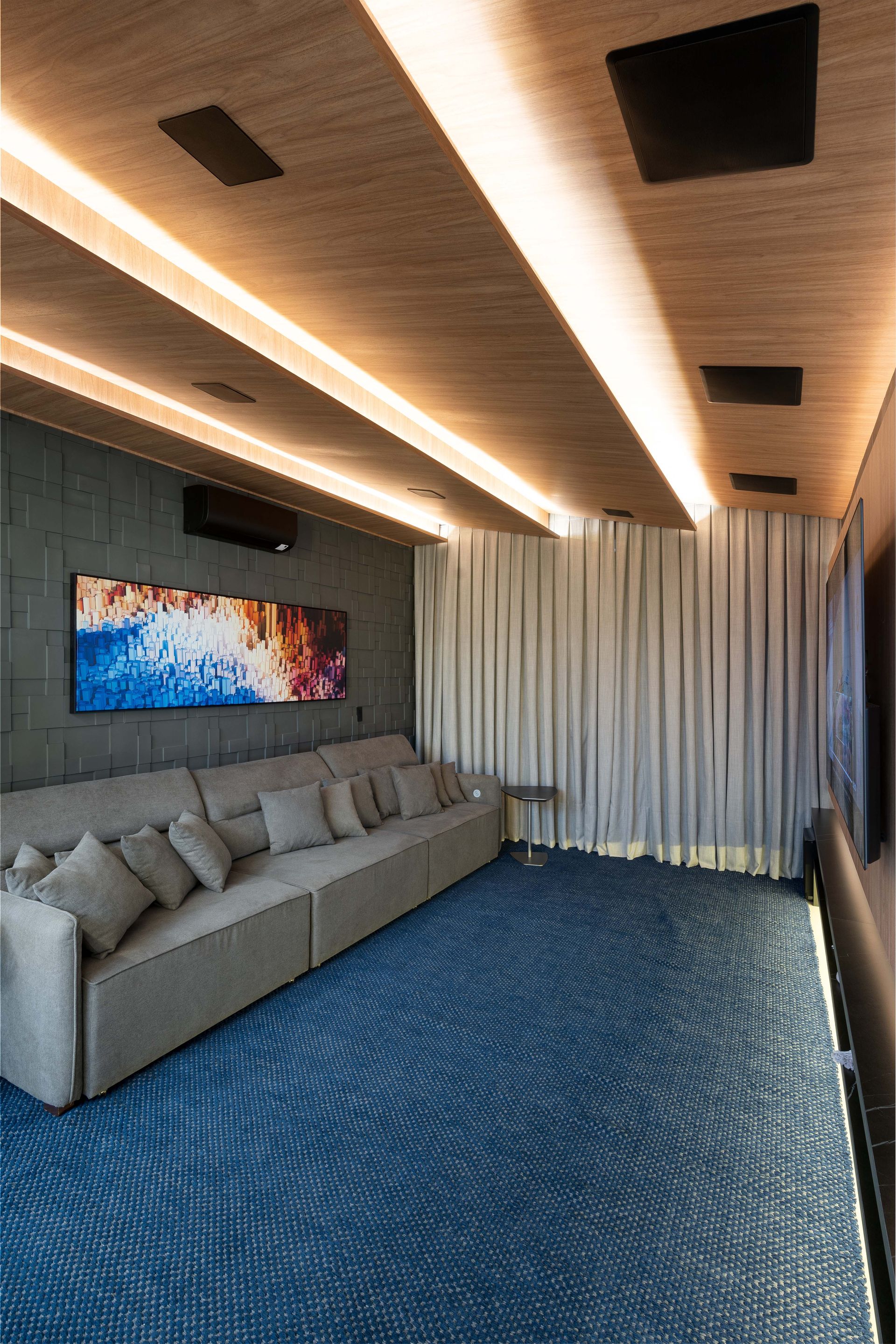 Home theater moderno com carpete azul, sofá cinza longo, arte e faixas de luz no teto.