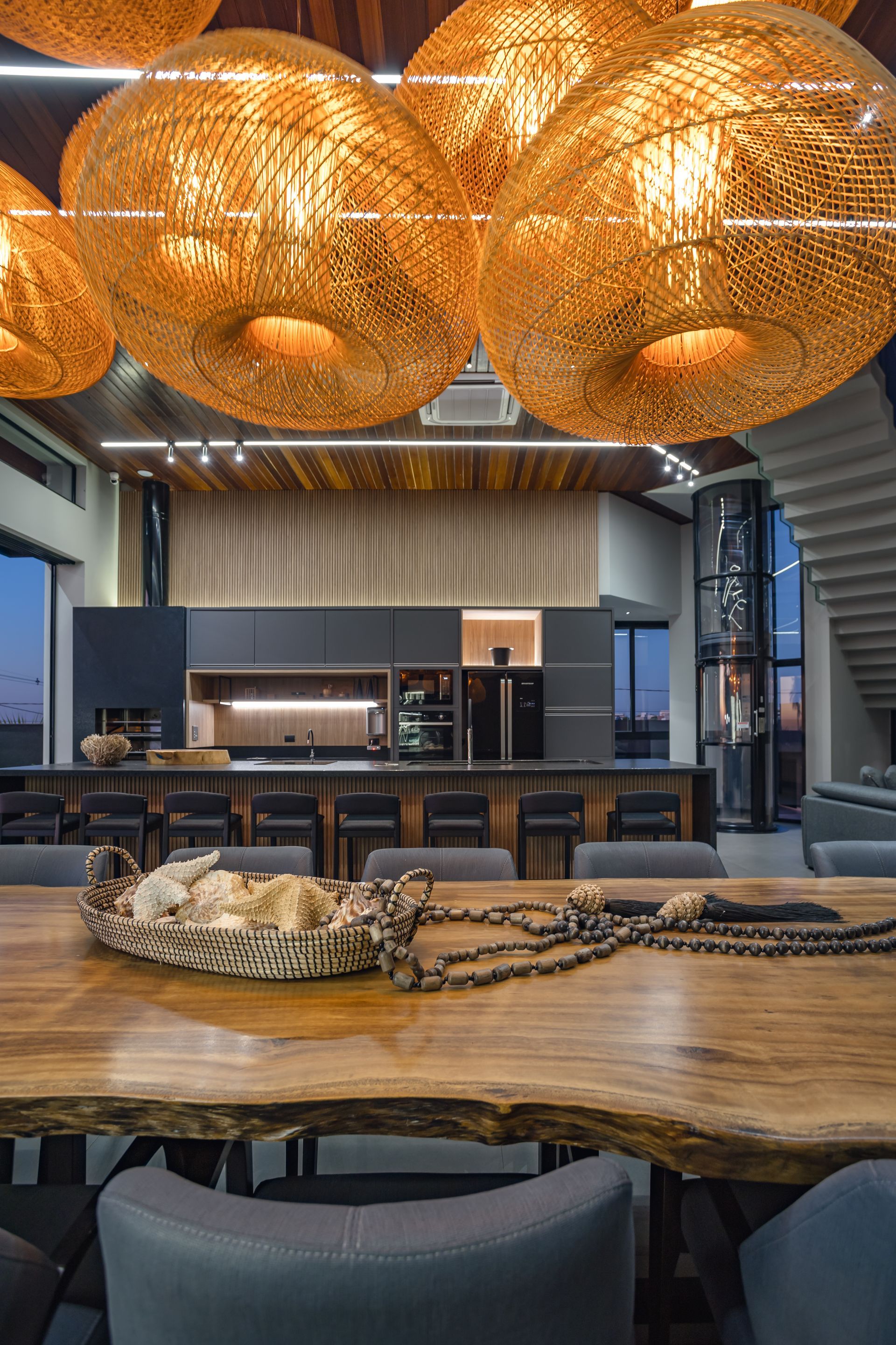 Sala de jantar elegante com mesa de madeira, luminárias pendentes de vime e cozinha ao fundo.